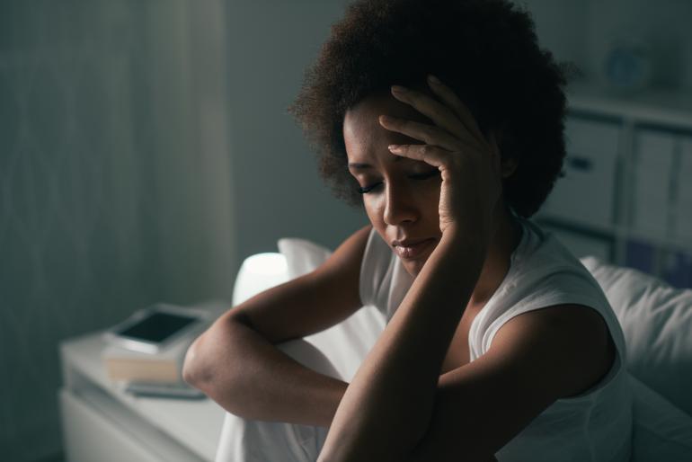 Early adult, thin Black woman with natural hair sitting in bed and holding her head with one hand.