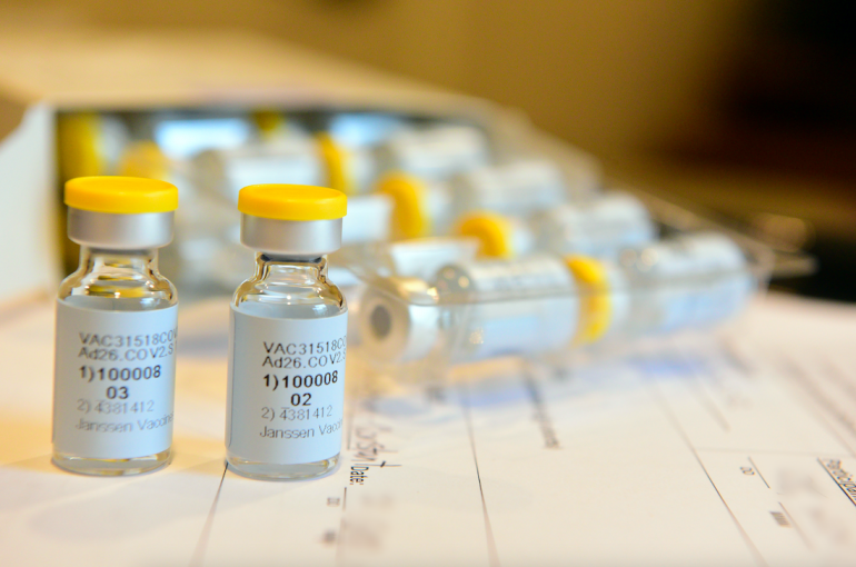 Vials of Janssen/Johnson & Johnson COVID-19 Vaccine Candidate sit on a desk. They are small clear vials with yellow lids and black and white labels.  