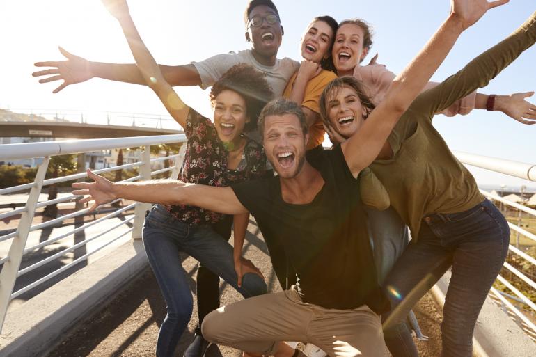 Group of smiling, multiracial young adults gather together with outstretched arms. They're wearing short sleeves and are standing on a pedestrian bridge on a sunny day.