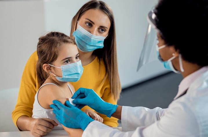A Black female doctor applies a bandaid to the arm of a white little girl who's sitting with her mother. We see the side and back of the doctor's head; she has short hair and is wearing a white coat, blue gloves, and blue surgical mask. The little girl has long medium-brown hair pulled back in a pony tail and is wearing a white tank top. Her mother sits with her arm around the little girl; she has long medium brown hair and is wearing a yellow long-sleeve shirt.