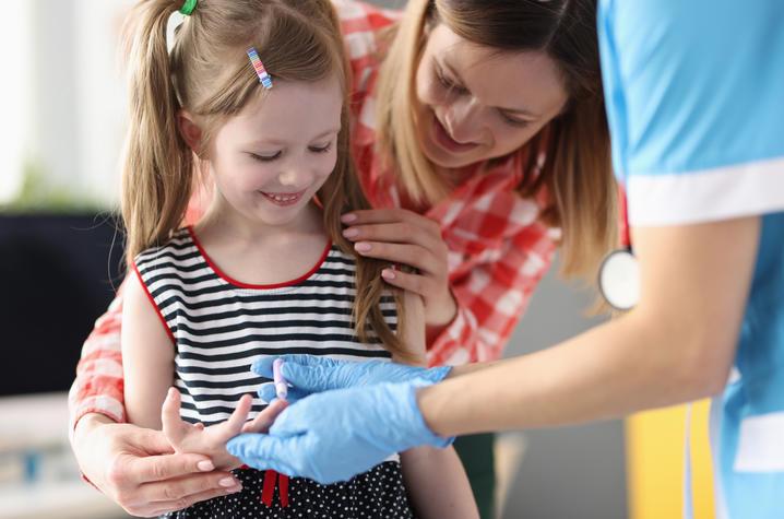 Stock image showing a child (female, white, with long hair pulled back in pig tails)  checking their blood sugar with a finger prick. Their mother and a health care provider assist. 