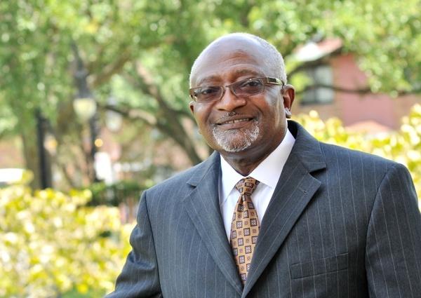 Dr. Robert Bullard in a dark gray suit stands smiling with trees out of focus behind him.