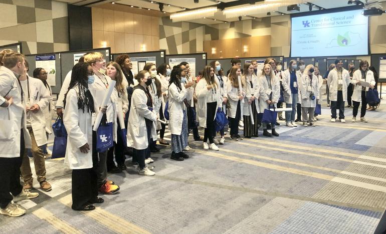 A group of about 50 high school students in white coats stand in a large conference room with research posters on board behind them. 