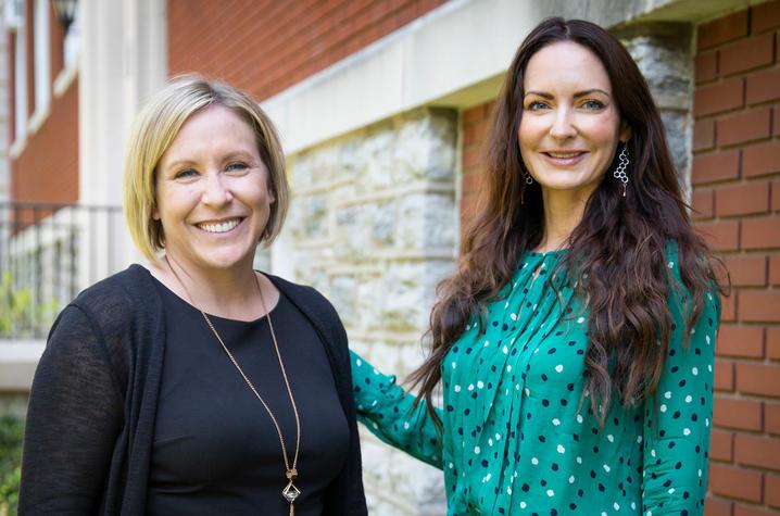 Waist-up photo of UK researchers Christal Badour, left, and Mairead Moloney standing outside a brick and stone building. Badour is on the left, a white woman with straight blond hair just past her chin. Moloney is on the right, a white woman with long, dark, wavy hair, wearing a teal blouse with dark and light blue dots. 