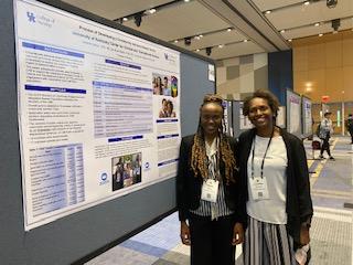 Hilda Okeyo, a PhD student in the UK College of Nursing, stands with Dr. Lovoria Williams. Hilda is a Black woman with long braided hair and she's wearing a suit. Lovoria is a Black woman wearing a white shirt, black sweater, and striped skirt. They're smiling and standing with a poster presentation pinned to a board. 