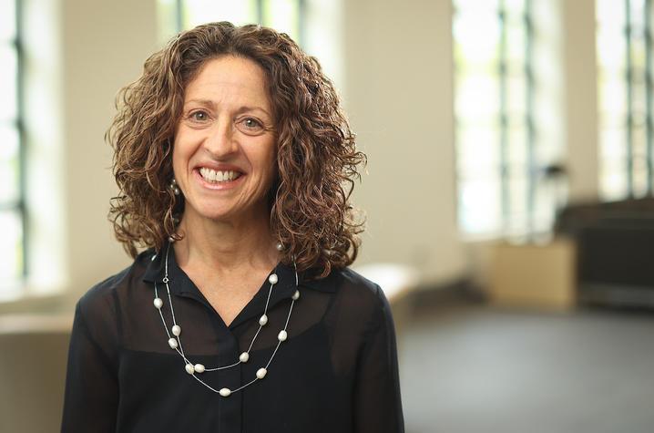 Torso headshot of Nancy Schoenberg, a middle-aged white woman with shoulder length curly hair. She's smiling at the camera, wearing a black collard blouse and a double drape necklace with intermittent pearls. 