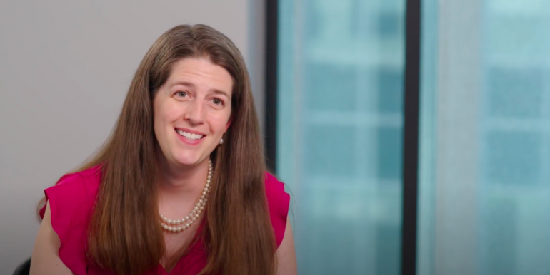 Dr. Mary Sheppard, a white woman in her mid 30s, smiles at the camera; she has long, thick, straight, light-brown hair and is wearing a pink short-sleeved blouse with a double strand of pearls. She's sitting in front of a light gray wall and a large window. 