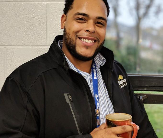 Robert Bell, a Black man in his late twenties, smiles at the camera. He's wearing a light blue striped collared shirt and a black jacket that's upzipped. He's standing in front of a window and holding a coffee mug. 