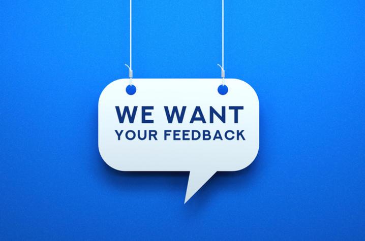 A white sign in the shape of a speech bubble says "We Want Your Feedback" in blue text. The sign hangs on white strings in front of a blue background. 