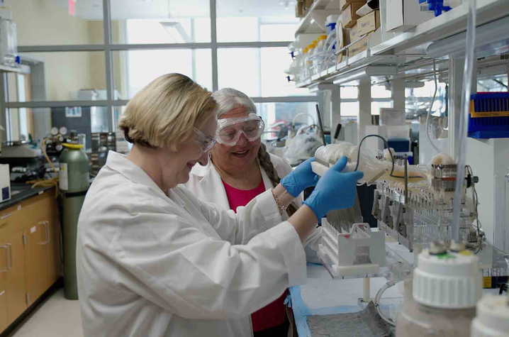 Two white women in their 50s or 60s, wearing white lab coats, blue medical gloves, and goggles, stand at the bench of a scientific laboratory; they're working with test tubes and equipment. 