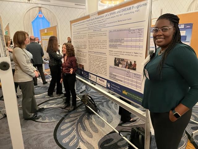 Brittany Smalls stands in the right side of the frame, next to a research poster she's presenting. She's a young Black woman with long hair, glasses, a smile, and wearing a dark green shirt and black trousers.