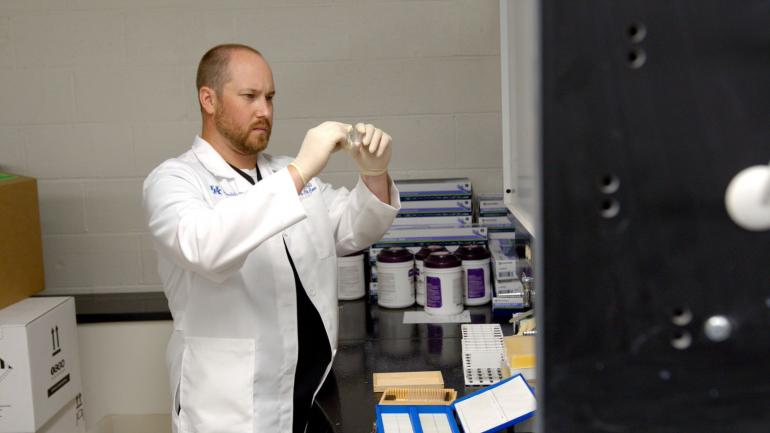 Kirby Mayer stands in a medical research lab; he's wearing a white lab coat and gloves and is holding up a microscope slide.