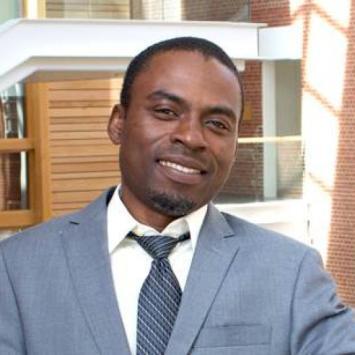 Headshot of Reuben Adatorwovor, PhD. He's standing outside of an academic building, wearing a gray tie, white collared shirt, and gray striped tie. He has very short black hair and a short black and gray beard on his chin. 