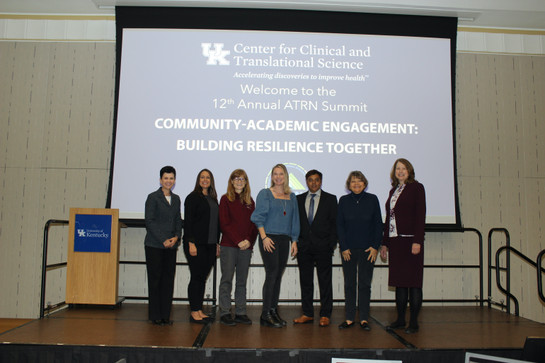 Seven ATRN leaders stand on a conference stage in front of a large projection screen. On the screen is a blue background with the following in white: logo for the UK Center for Clinical and Translational Science, below which is the text "Welcome to the 12th Annual ATRN Summit, Community and Academic Engagement: Building Resilience Together
