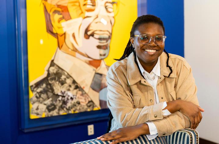 Princess Magor Agbozo, an undergraduate student in public health, stands in front of a large, bright yellow portrait collage of Lyman T.  Johnson. Agbozo is a young Black woman wearing a beige corduroy jacket with a white collared shirt underneath. She's wearing clear-rimmed glasses, her long twists pulled back in a ponytail, and she's smiling at the camera while leaning on the back of a striped wingback chair. 