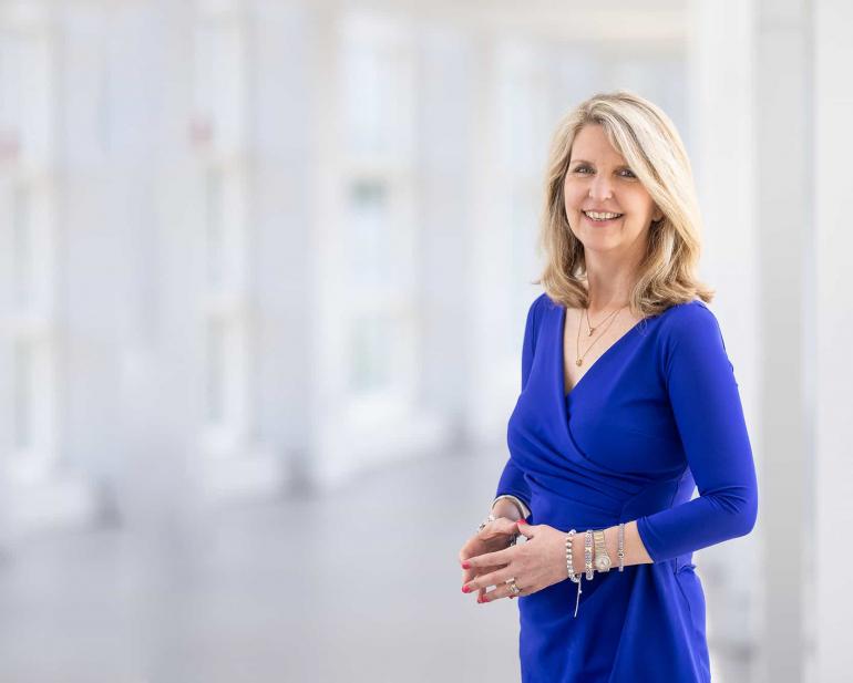 Dr. Susan Smyth, a white woman with blond, shoulder length hair, stands in a white room with floor to ceiling windows behind her. She's wearing a royal blue v-neck dress and several bracelets.