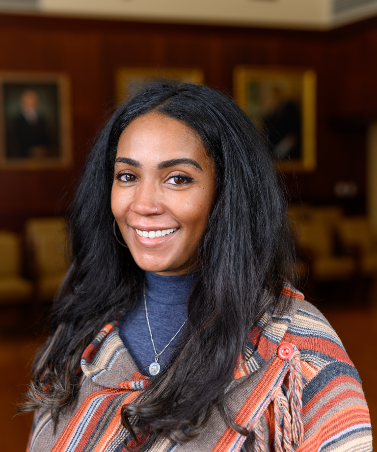 Headshot of Laneshia Conner, PhD, a Black woman in her 30s with long straight hair; she's smiling at the camera wearing salmon/blue/gray striped sweater and a blue turtle neck. In the blurred background is a maroon wall with large portraits.