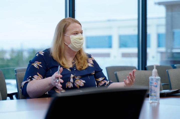 Alison Gibson, a white woman in her 30s, sits at a conference table with a wall of windows behind her. She has shoulder-length blonde hair, wearing a yellow procedure mask, and a navy blouse with big pink flowers. She's gesturing with her hands. 