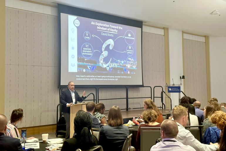 David Butts, a middle-aged Black man in a suit, speaks from a podium to a room full of people. Behind him is a projection screen with a slide that says "Exploration towards a Mindset of Equity."