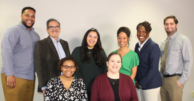 Photo of 8 adults of mixed races and skin tones. 6 adults are standing and two are sitting. They're in front of a white wall. Everyone is dressed in business casual clothes and smiling at the camera.