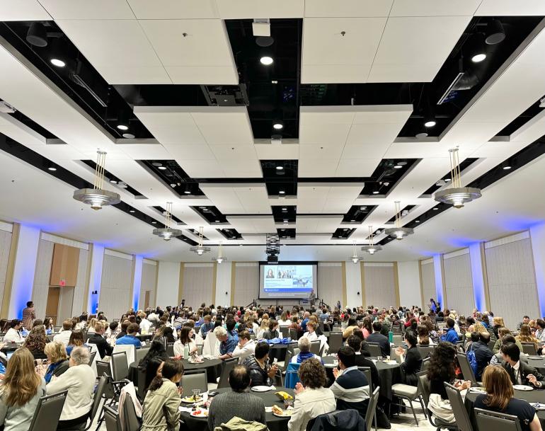 Photo of a very large room filled with hundreds of conference participants sitting at round tables. The view is from the back of the room, looking to the front of the room where a large project screen seems small in the distance.