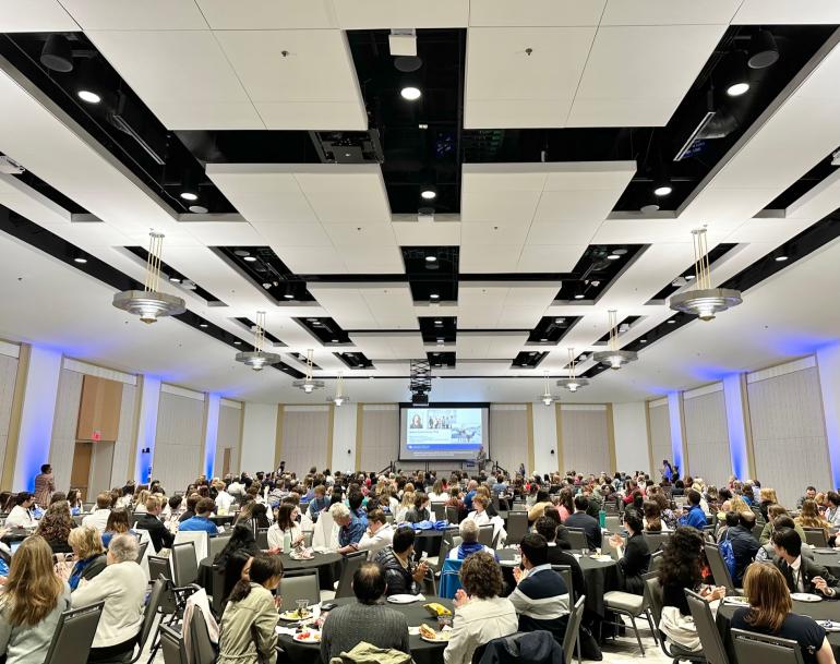 Photo of a very large room filled with hundreds of conference participants sitting at round tables. The view is from the back of the room, looking to the front of the room where a large project screen seems small in the distance. 