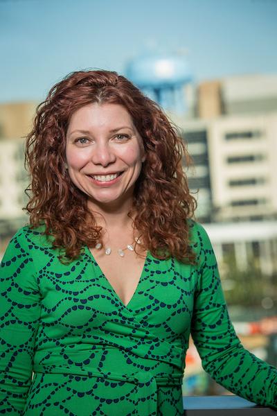 Photo of Michelle Hernandez, a middle aged woman with light skin and curly reddish-brown hair past her shoulders. She's wearing a v-neck green dress with dark blue pattern and is standing outside. 