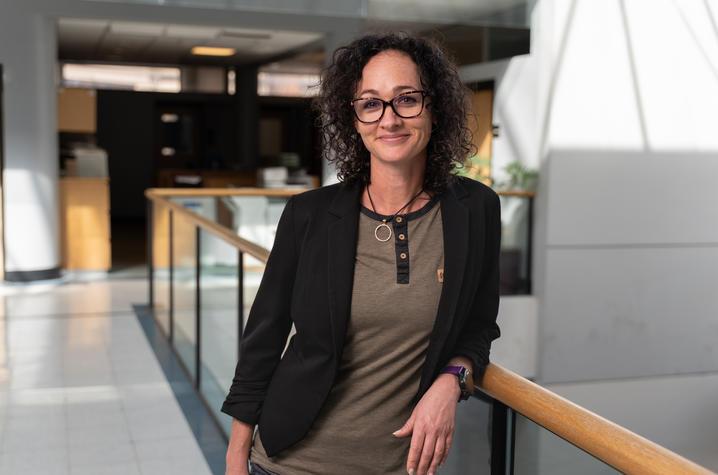 Photo of Sally Ellingson, PhD. She's a white woman in her 30s with very curly dark brown hair down to her shoulders and dark-rimmed glasses. She's wearing a dark brown blazer and a light brown shirt with dark trim. She's standing in a brightly lit building and leaning against a wooden guard rail. She's smiling at the camera.
