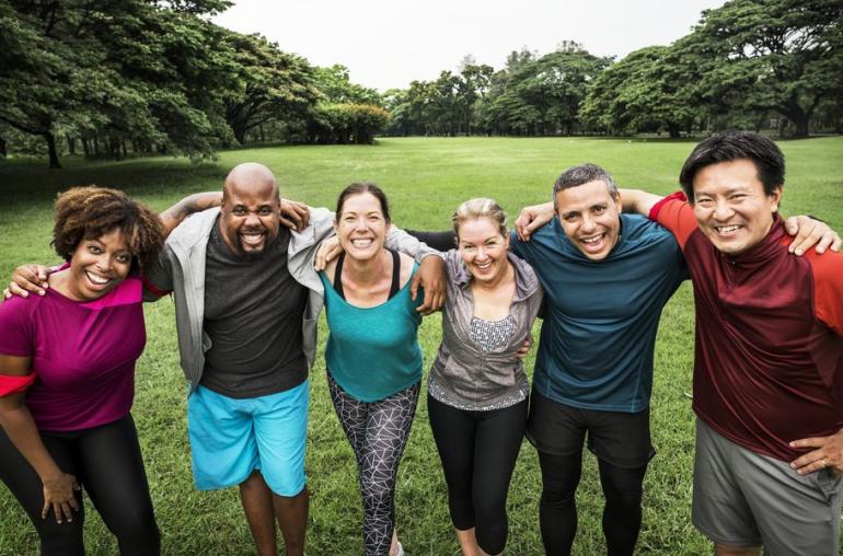 Group of six adults of various ages stand in a park-like setting with their arms around each others' shoulders. They're smiling at the camera and wearing workout clothes. 
