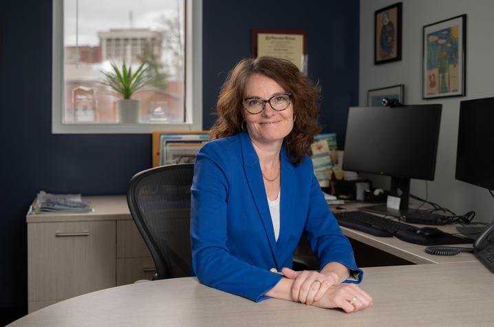Portrait of Erin Haynes sitting at her desk in her office. She's wearing a royal blue blazer with a white shirt beneath and her hands are folded together on the table. She has reddish-brown wavy hair just past her shoulders and she's wearing glasses. Behind her is a dark blue wall with a window, and to the right  are her computer monitors and framed pictures on a white wall. 