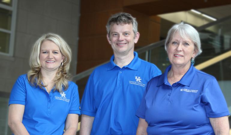 The three members of the CCTS Biobank team stand together wearing matching blue polo shirts with the UK CCTS logo embroidered on the left chest. The woman on the left is white with blond hair past her shoulders; the man in the middle is white with short brown and gray hair, and the woman on the right is white with short gray hair. They're smiling at the camera. 