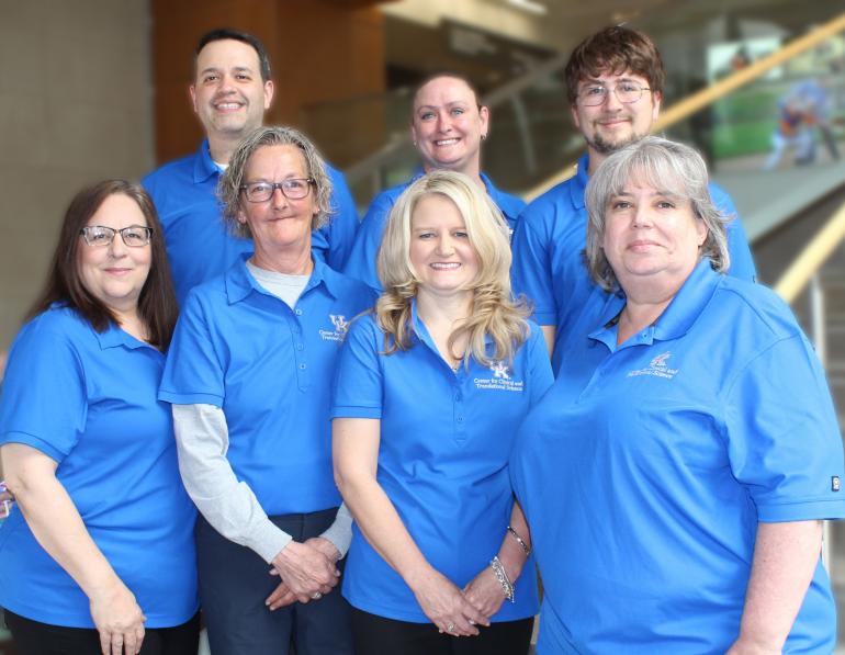 The seven members of the CCTS Regulatory team stand together, all wearing bright blue polo shirts with the CCTS logo on the left chest. Three women stand in front, with two men and a woman standing behind them, slightly elevated on a step. 