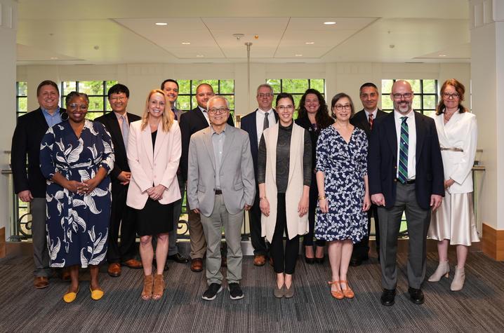 A diverse group of 16 faculty members stand together. They are all dressed professionally and behind them is a wall of windows. 