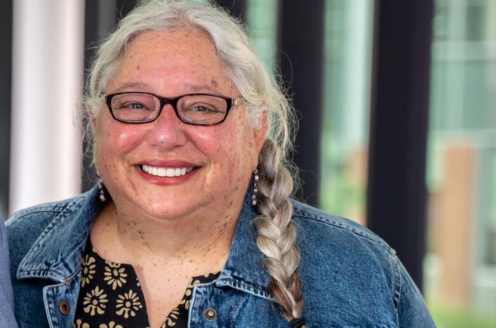 Linda Dwoskin is a white woman with long gray hair in a braid over her shoulder. She's smiling at the camera, wearing black-rimmed glasses, a black blouse with with flowers, and a denim jacket. A wall of windows is behind her. 