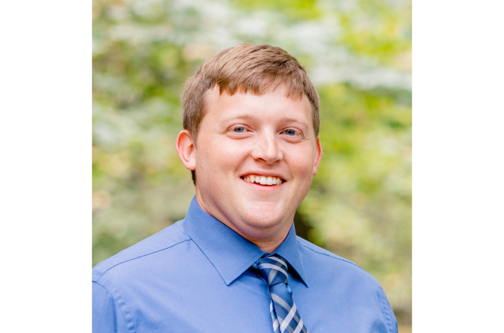 Headshot of Matthew Coleman, a young white man with short, light brown hair. He's outdoors, with trees blurred in the background, and he's wearing a light blue collared shirt with a blue and gray striped tie. 