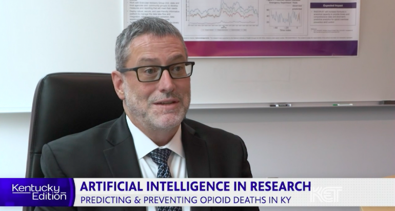 Screenshot of interview with Jeff Talbert, PhD. He's a white, middle-aged man sitting in an office chair wearing a gray suit, white shirt, and dark tie with black-rimmed rectangular glasses and short gray hair. There's a white board behind him and the bottom of a research poster is visible. At the bottom of the screen is a text banner that says "Kentucky Edition. Artificial Intelligence in Research: Predicting & Preventing Opioid Deaths in KY"