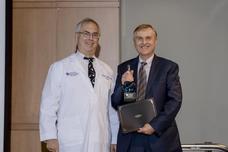 Dr. Philip Kern receives the 2023 Willard award. He's wearing a dark suit, holding a small statue and a certificate. Beside him is a man in a white medical coat. They're both smiling. 