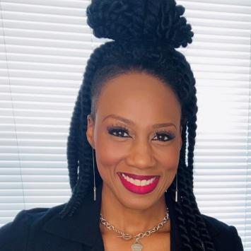 Headshot of Ketrell McWhorter, a young Black woman with shoulder-length twists, half pulled up into a high bun. She's smiling at the camera, wearing red lipstick, a black suit jacket, and there's a window with blinds behind her. 