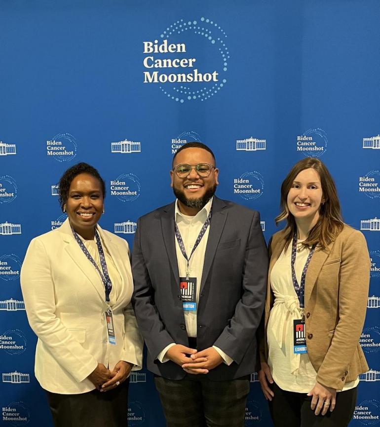 Three adults stand in front of a royal blue backdrop that says "Biden Cancer Moonshot" at the top. On the left is a Black woman wearing a cream colored blazer and dark trousers; in the center is a Black man with glasses, gray blazer, cream shirt, and plaid trousers; and on the left is a white woman in a tan blazer, cream blouse, and dark trousers. They're each smiling and wearing an event lanyard. 