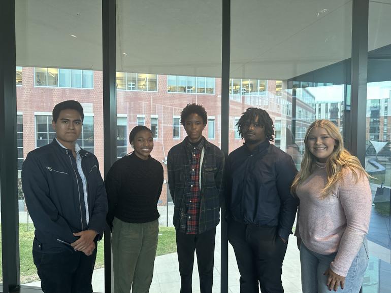 Five undergradute students stand side by side in front of a floor-to-ceiling window.