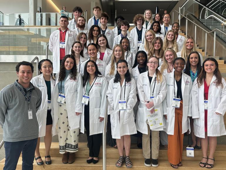 33 students from Frederick Douglass High School, all wearing white lab coats, and two of their teachers stand on the steps in Central Bank Center. 