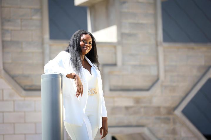 A young Black woman dressed in a white suit with gold belt stands and leans her right elbow on a gray bollard. She has chest-length black hair and is smiling at the camera. In the background is a very large stone sculpture of the UK inisignia. 