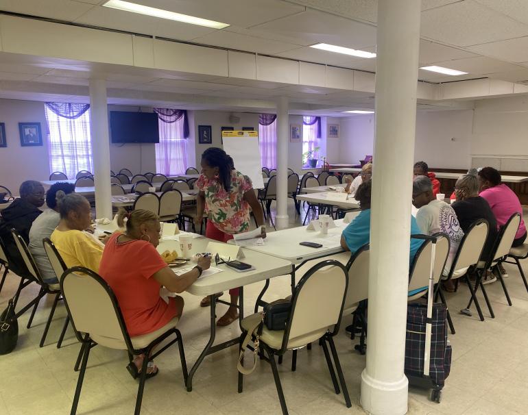 A focus group meets to discuss health information handouts. Eleven Black adults sit at long folding tables arranged in a U-shape. In the center, a Black woman leading the group leans over one of the tables to talk to a participant. 