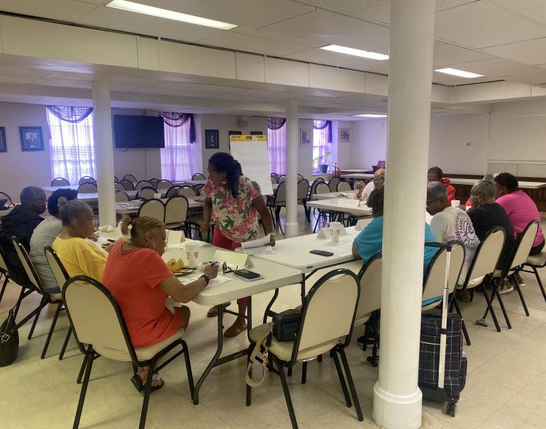 A focus group meets to discuss health information handouts. Eleven Black adults sit at long folding tables arranged in a U-shape. In the center, a Black woman leading the group leans over one of the tables to talk to a participant.
