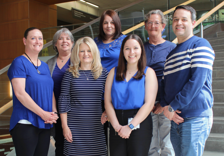Group shot of seven white adults standing on sunny interior stairs. They're all wearing shades of blue and smiling at the camera. 