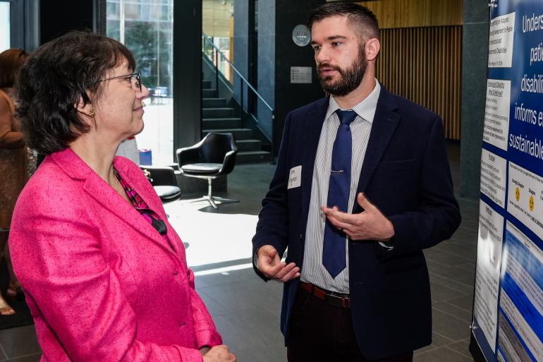 A young white man in a dark blue suit and navy tie presents a research poster to NIH director Monica Bertagnolli, a middle-aged white woman wearing a pink suit. 