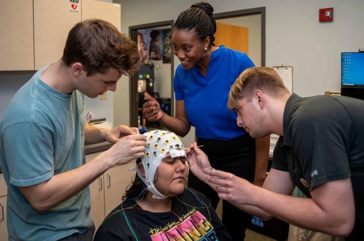 Dr. Lauren Whitehurst, a young Black woman with her hair in a bun, stands with two of her students who are setting up an electrode cap on the head of a research participant.