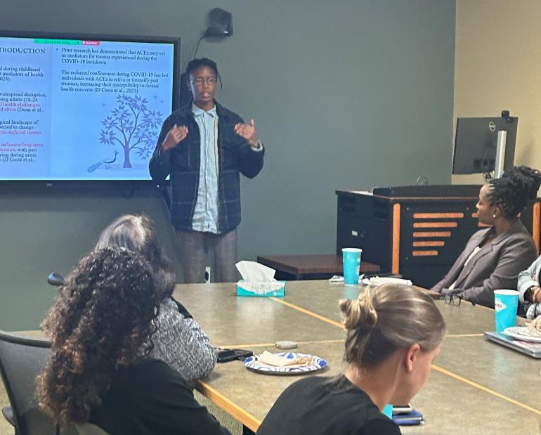 Photo of a young Black man giving a presentation to several people at a conference table. There's a digital screen behind him, displaying information about his research, and he's gesturing with his hands as he speaks.