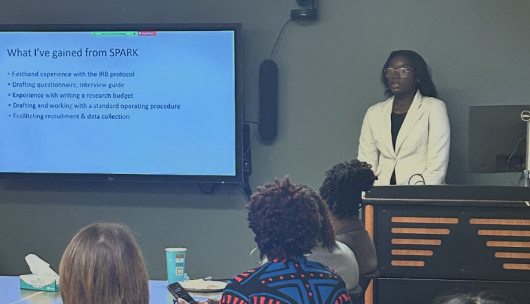 A young Black woman gives a research presentation to a several people sitting around a conference table. She is standing at the front of the room next to a digital screen displaying her presenation. She's wearing a white suit jacket and glasses, with long black hair.
