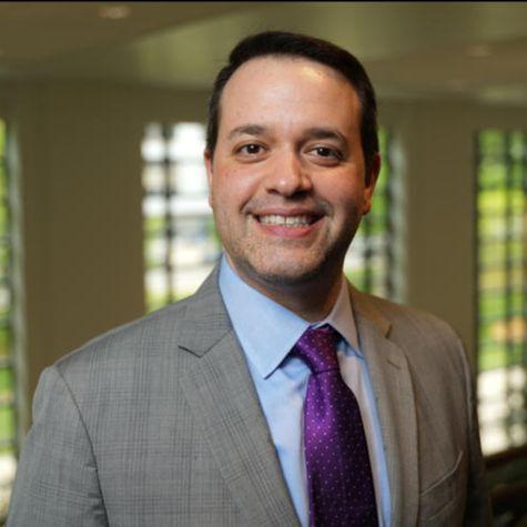 Headshot of Bill Stoops, a middle-aged white man whit short dark hair. He's smiling at the camera, wearing a light gray suit, light blue shirt, and dark purple tie. There's a wall of tall windows behind him. 